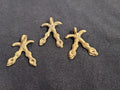Three small brass Akofena pendants shaped as crossed swords, each with a hanging loop, arranged on dark fabric