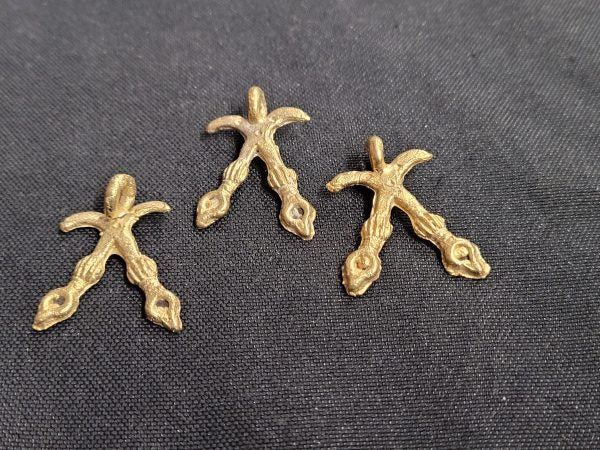 Three small brass Akofena pendants shaped as crossed swords, each with a hanging loop, arranged on dark fabric