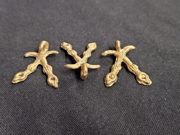 Three brass crossed-swords Akofena pendants arranged in a row on dark fabric, showing variations in their handmade shapes
