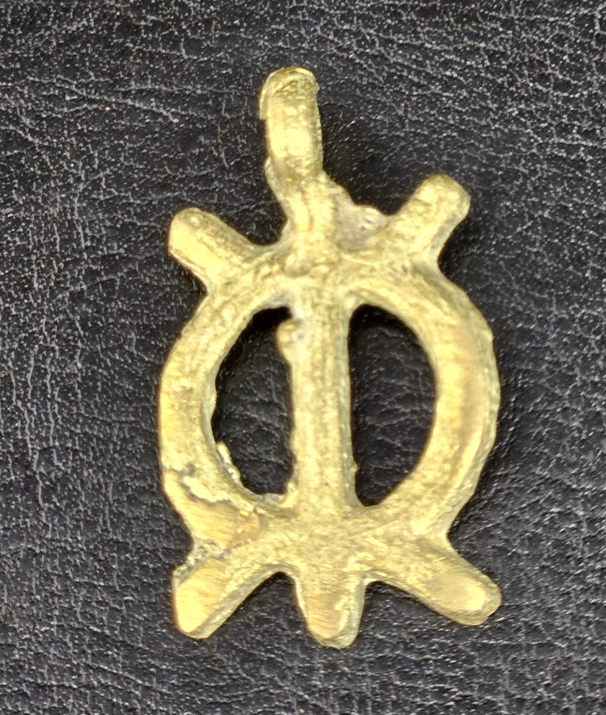 Small brass Wawa Aba pendant with an oval openwork center divided by a vertical bar, four projecting points, and a top hanging loop on dark fabric