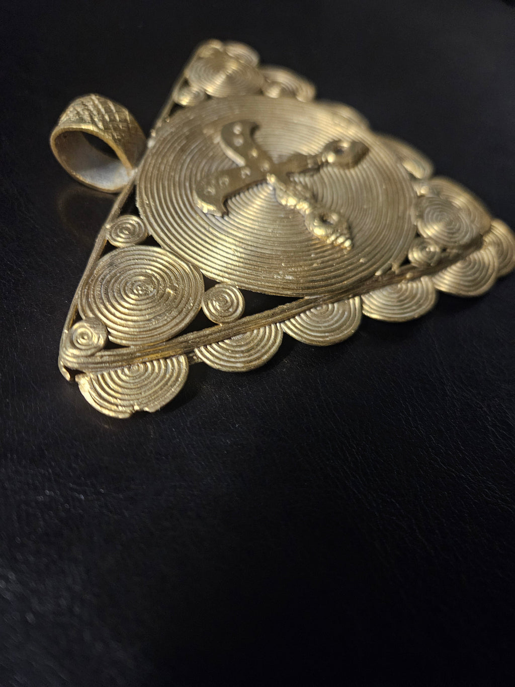 Angled view of the triangular brass Akofena pendant showing its raised crossed-swords motif, spiral detailing, and hanging loop.