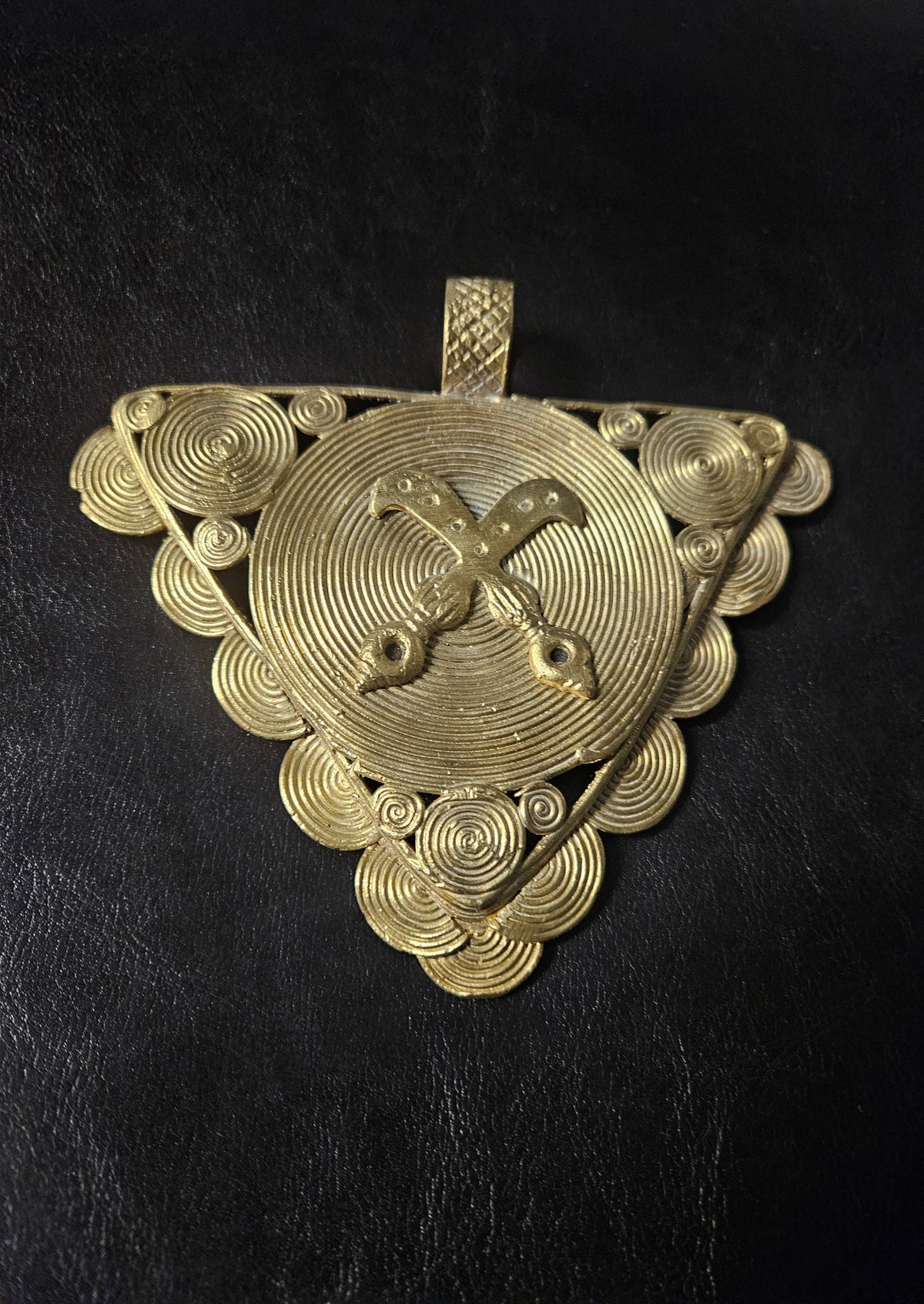 Triangular brass Akofena pendant with raised crossed swords over a large spiral disc, smaller spiral discs around the border, and a textured top hanging loop