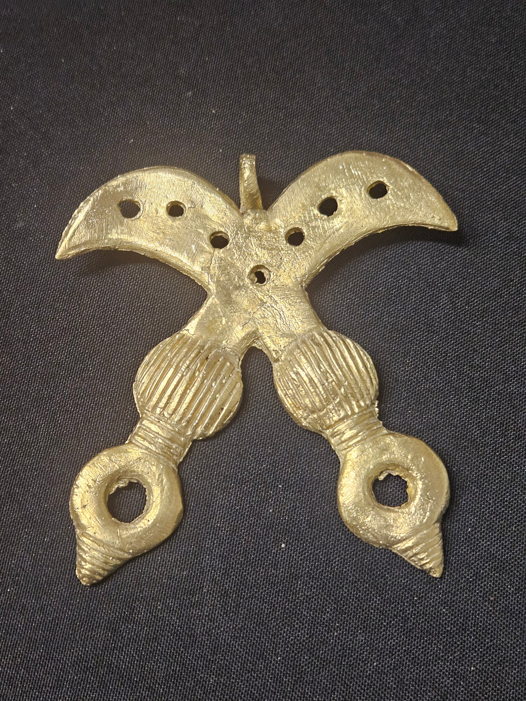 Brass Akofena pendant shaped as crossed swords, with dotted curved blades, patterned handles, and a top hanging loop on dark fabric