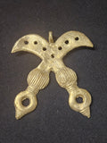 Brass Akofena pendant shaped as crossed swords, with dotted curved blades, patterned handles, and a top hanging loop on dark fabric