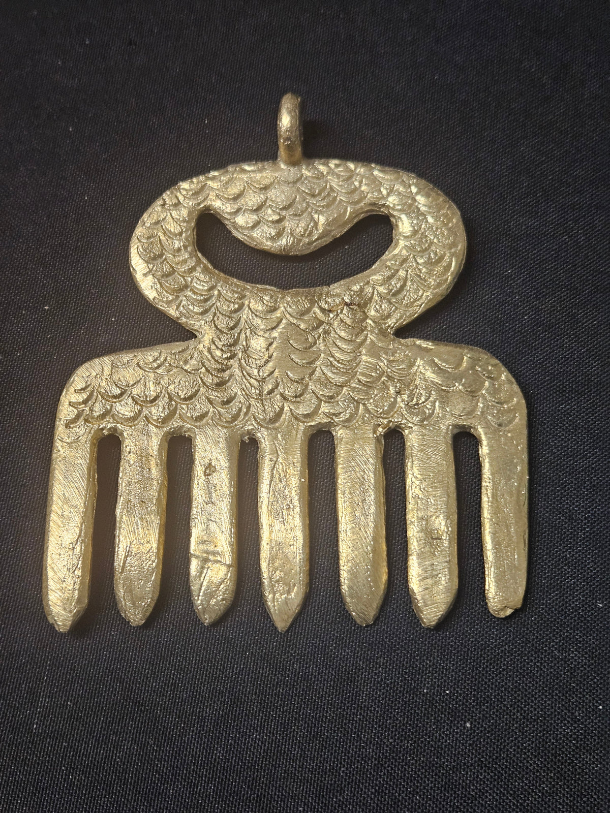 Brass Duafe pendant shaped like a comb, with an oval openwork handle, patterned surface, and a top hanging loop on dark fabric