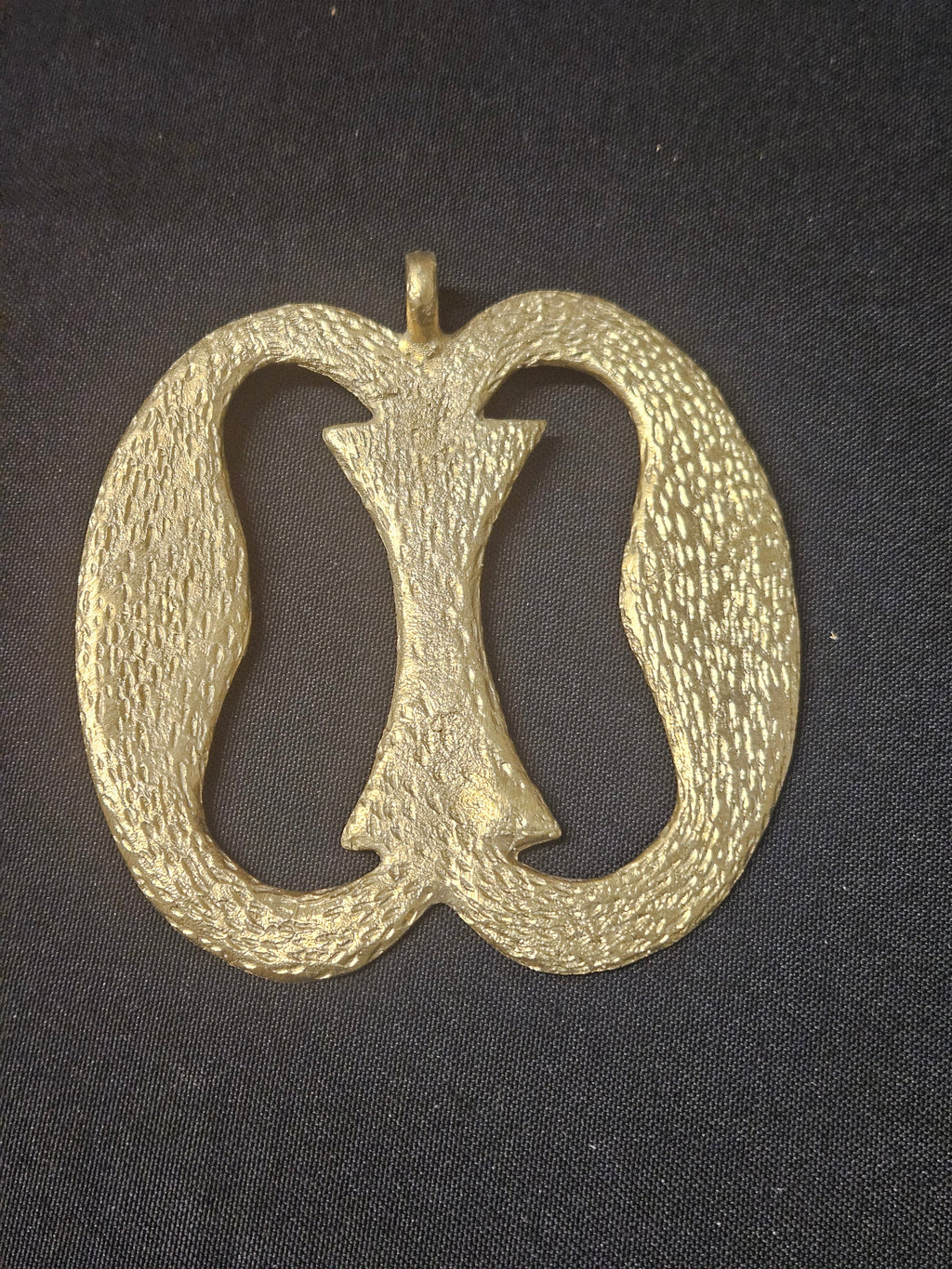 Brass Nkonsonkonson pendant with two elongated open loops joined by a central bar, a textured surface, and a top hanging loop on dark fabric