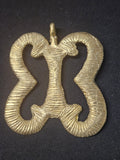 Brass Pempamsie pendant with four inward-curving open sections joined by a central bar, grooved surface details, and a top hanging loop on dark fabric