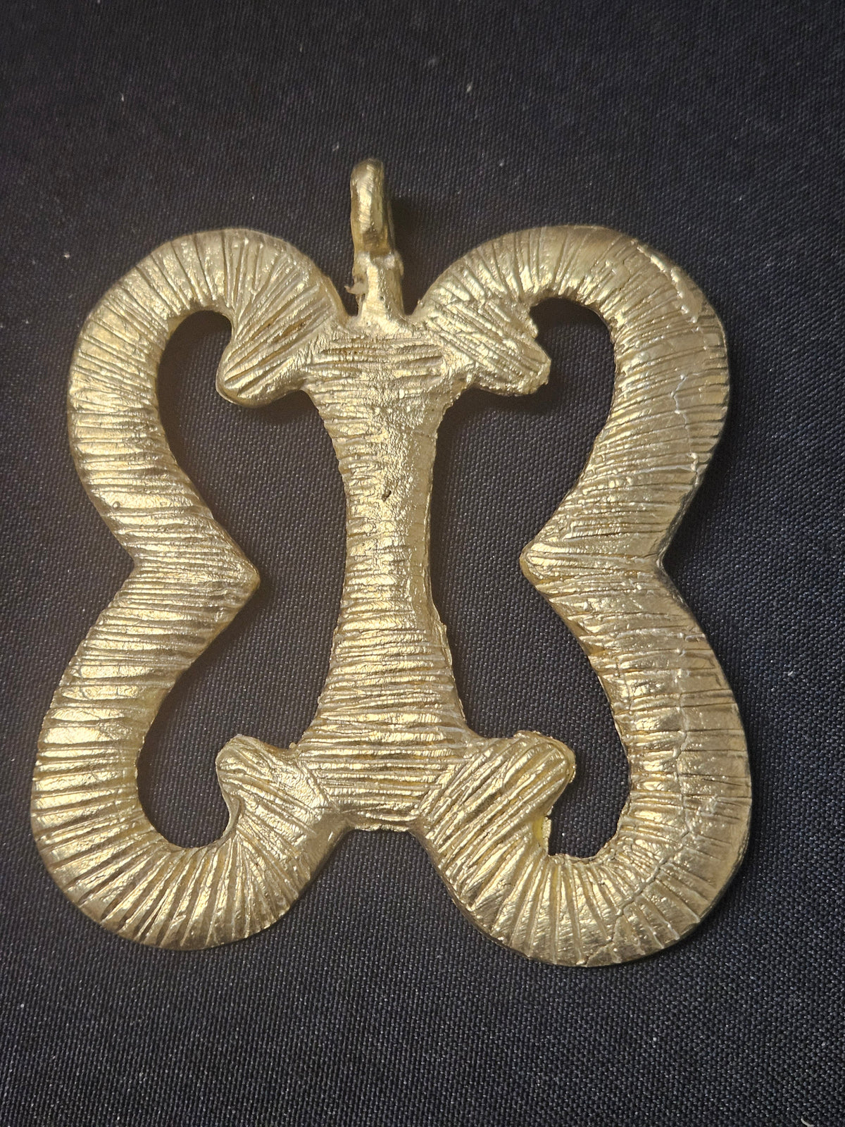 Brass Pempamsie pendant with four inward-curving open sections joined by a central bar, grooved surface details, and a top hanging loop on dark fabric