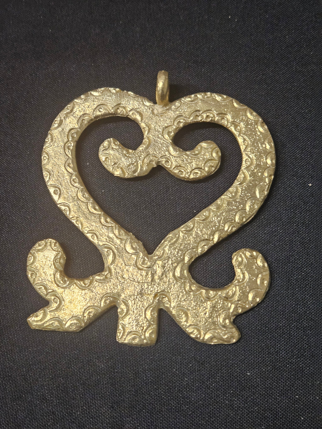 Brass Sankofa pendant in an open heart-shaped design with inward curls, textured surface patterns, and a top hanging loop on dark fabric