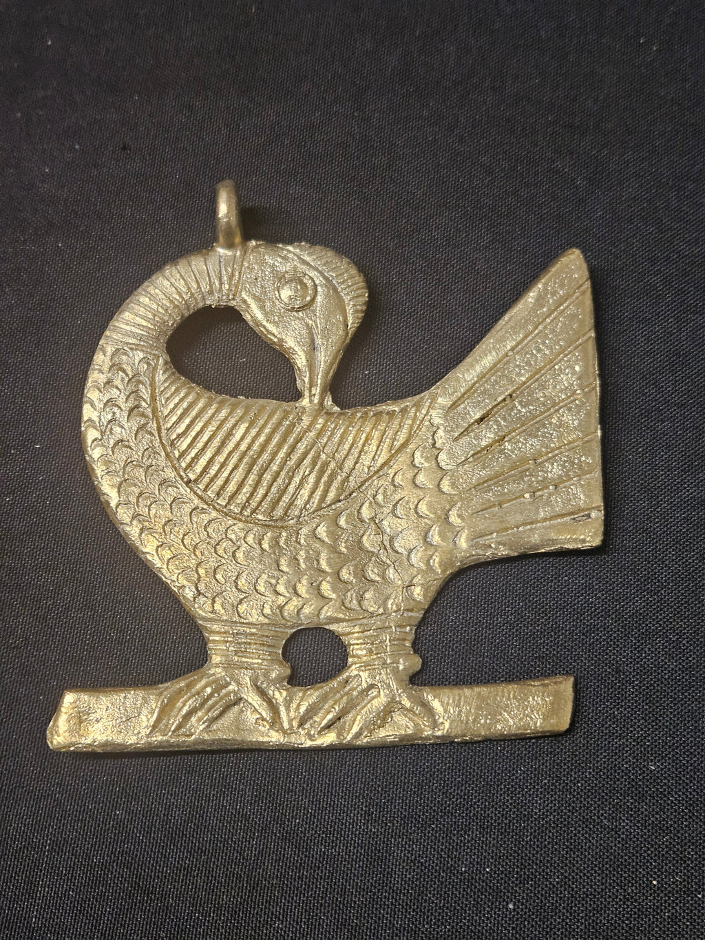 Brass Sankofa bird pendant with its head turned backward, patterned feathers, a fan-shaped tail, and a top hanging loop on dark fabric