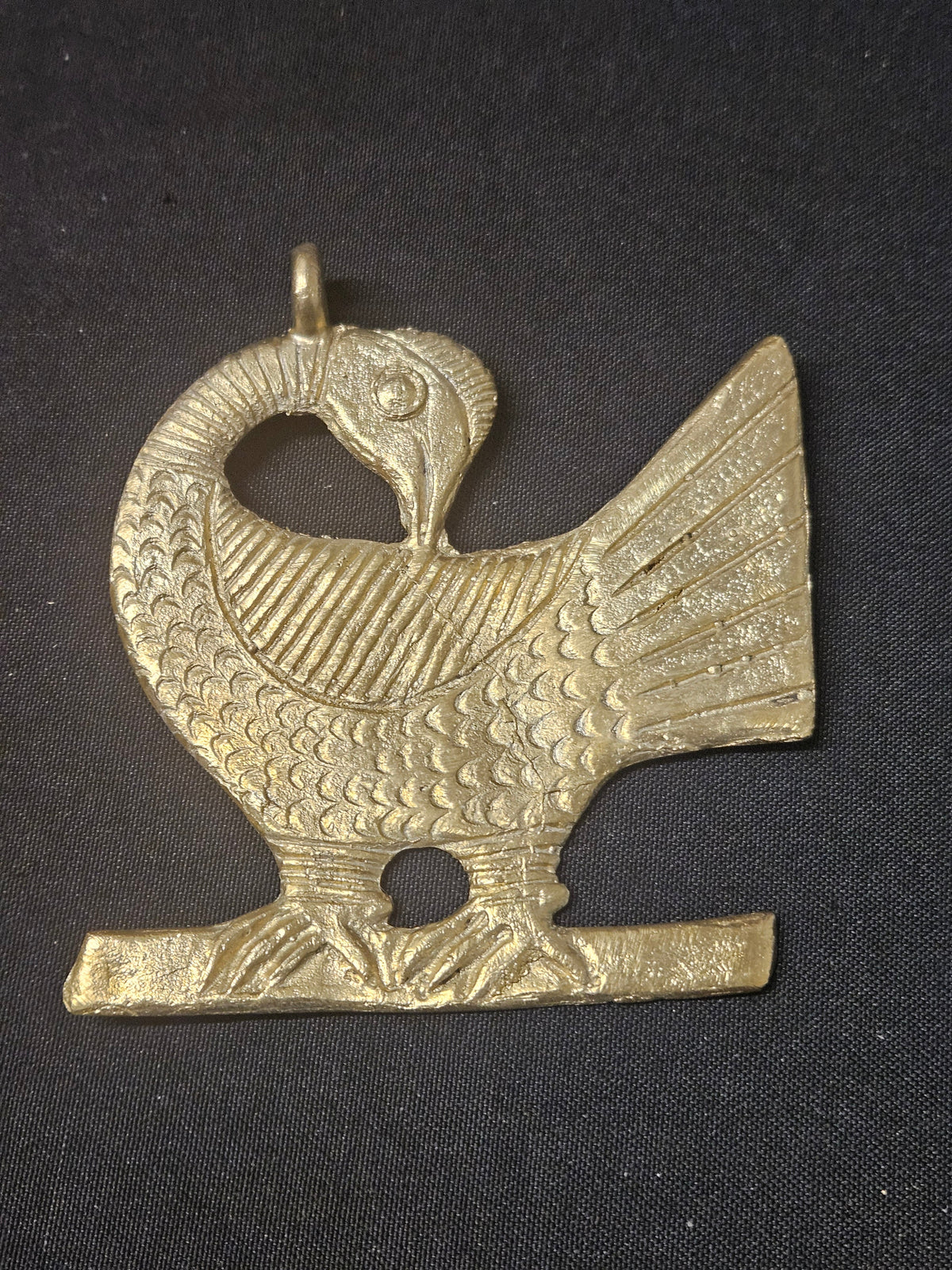 Brass Sankofa bird pendant with its head turned backward, patterned feathers, a fan-shaped tail, and a top hanging loop on dark fabric