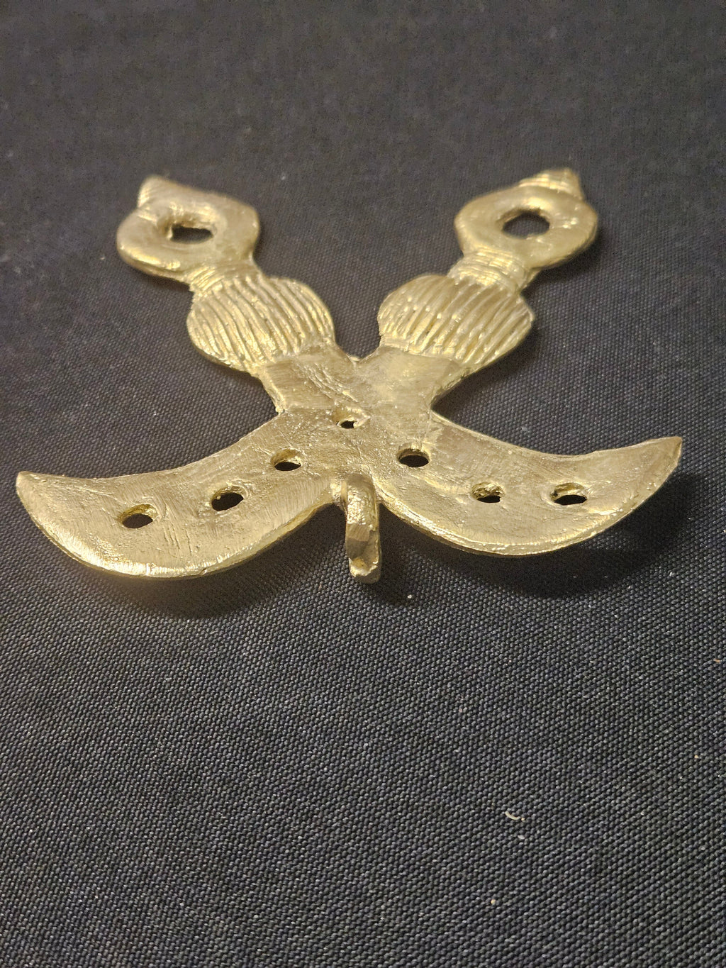 Brass crossed-swords Akofena pendant rotated with the handles at the top, showing its curved blades and decorative openings on dark fabric