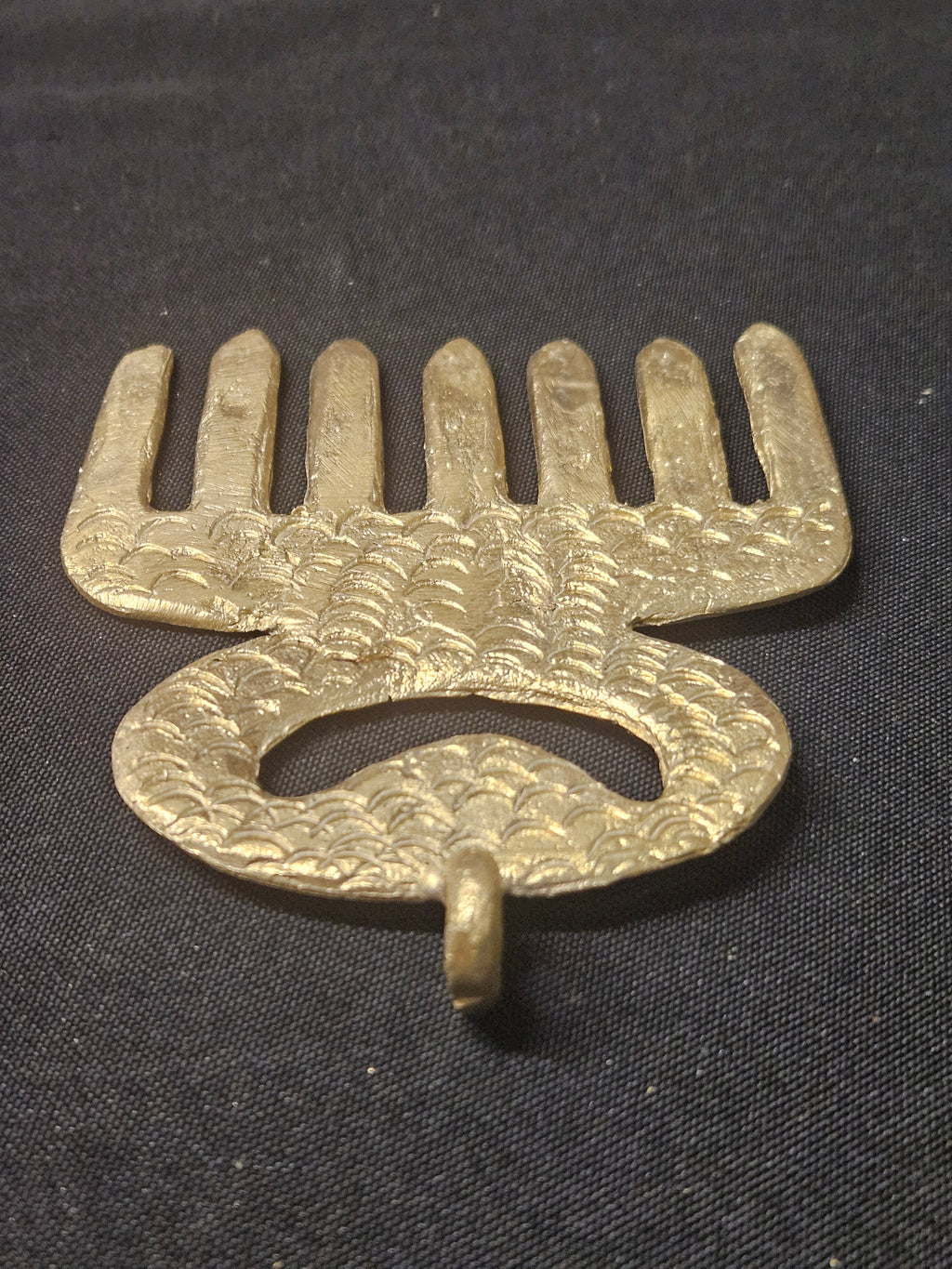 Angled view of the brass Duafe comb-shaped pendant, rotated with its teeth at the top, showing the openwork handle and thickness on dark fabric