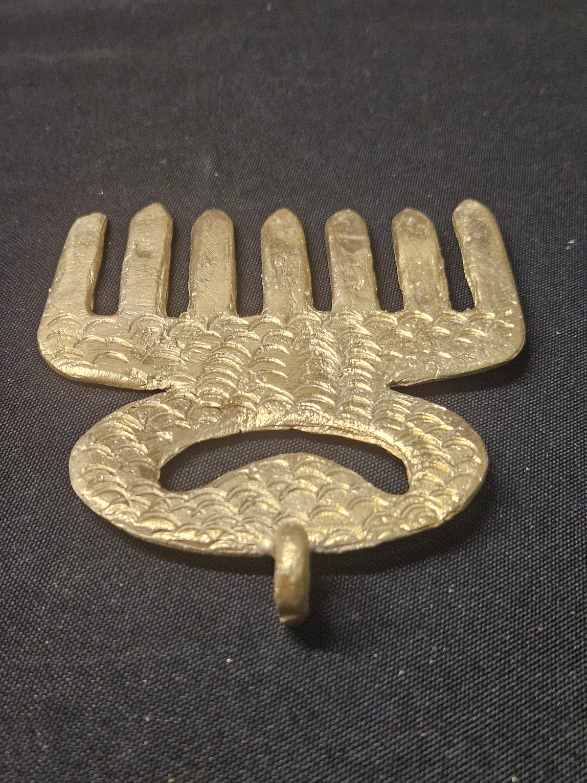 Angled view of the brass Duafe comb-shaped pendant, rotated with its teeth at the top, showing the openwork handle and thickness on dark fabric