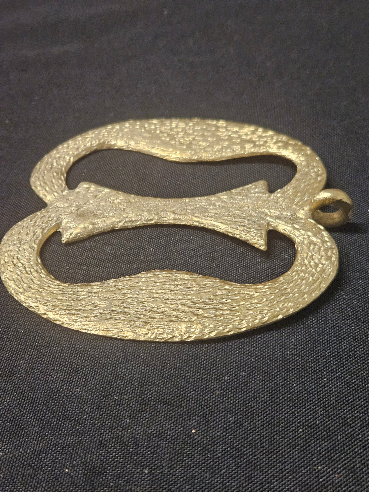 Low angled view of the brass Nkonsonkonson pendant showing its two open loops, central connecting bar, and thickness on dark fabric