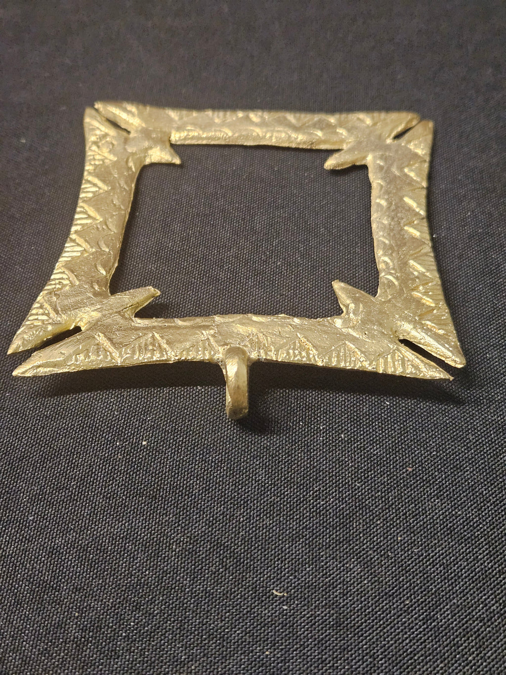 Angled view of the brass Fihankra pendant with its hanging loop at the bottom, showing the square opening, pointed corners, and thickness on dark fabric