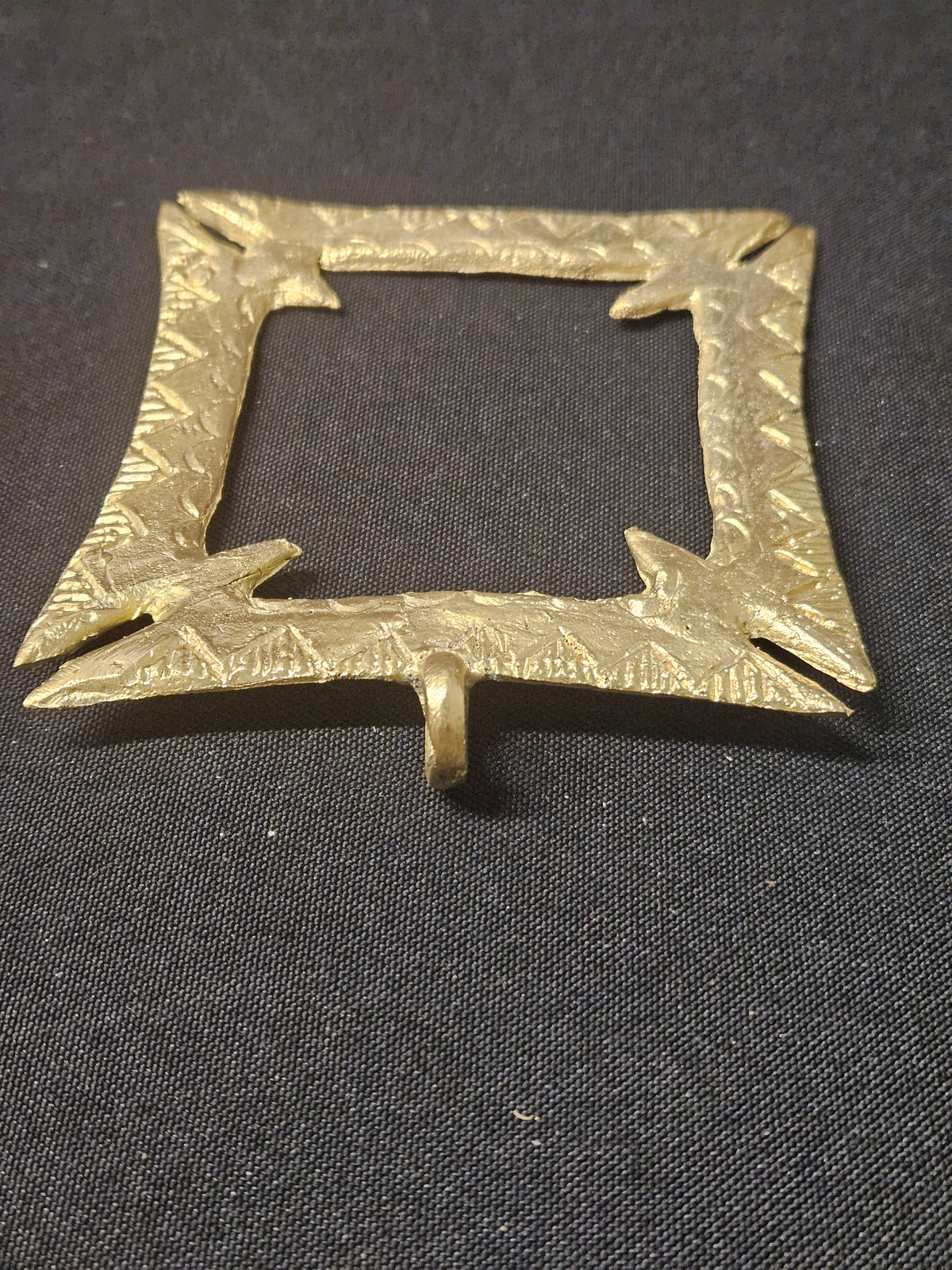 Angled view of the brass Fihankra pendant with its hanging loop at the bottom, showing the square opening, pointed corners, and thickness on dark fabric