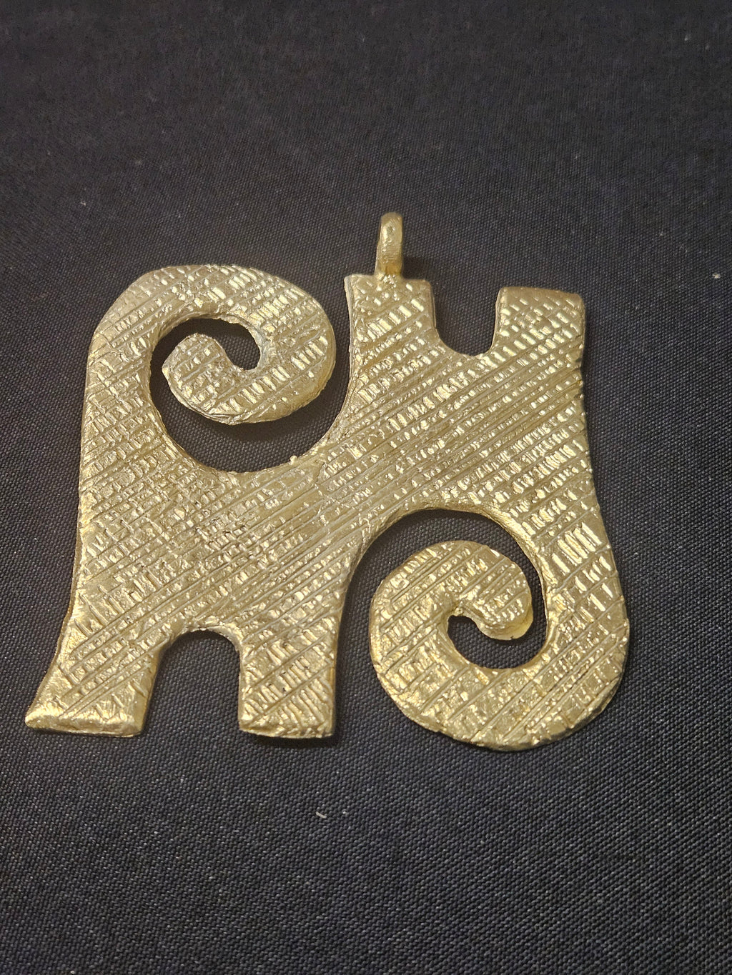 Brass Kwaatakye Atiko pendant with two diagonal spiral curls, a textured central section, edge notches, and a top hanging loop on dark fabric