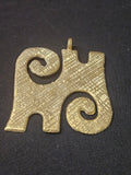 Brass Kwaatakye Atiko pendant with two diagonal spiral curls, a textured central section, edge notches, and a top hanging loop on dark fabric