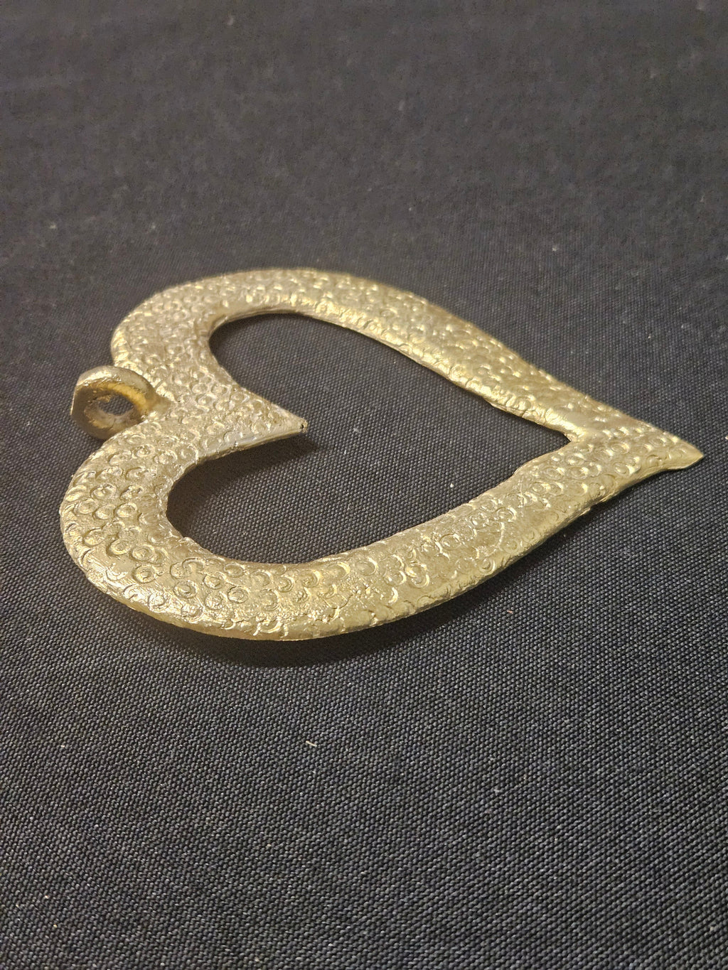 Brass open-heart Akoma pendant lying sideways on dark fabric, showing its heart-shaped outline and hanging loop