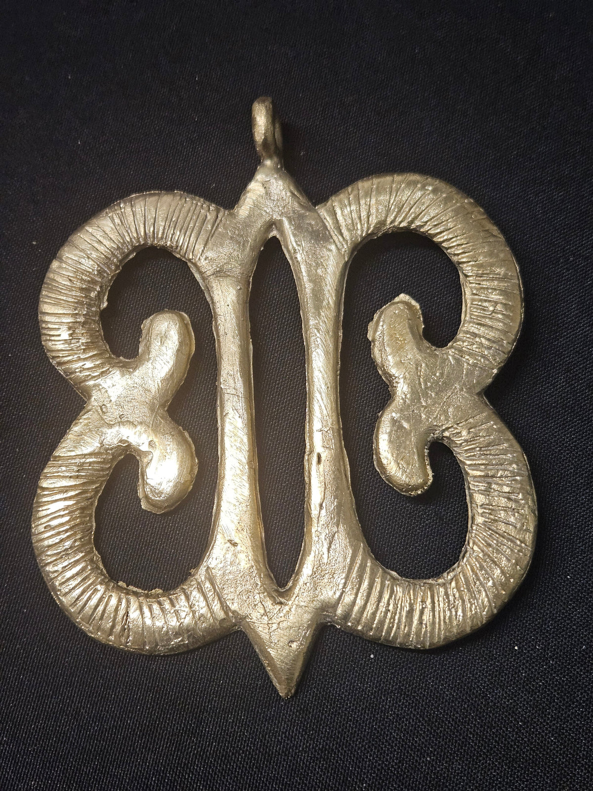 Brass Hye Won Hye pendant with four curled open loops around an elongated central opening, grooved surface details, and a top hanging loop on dark fabric