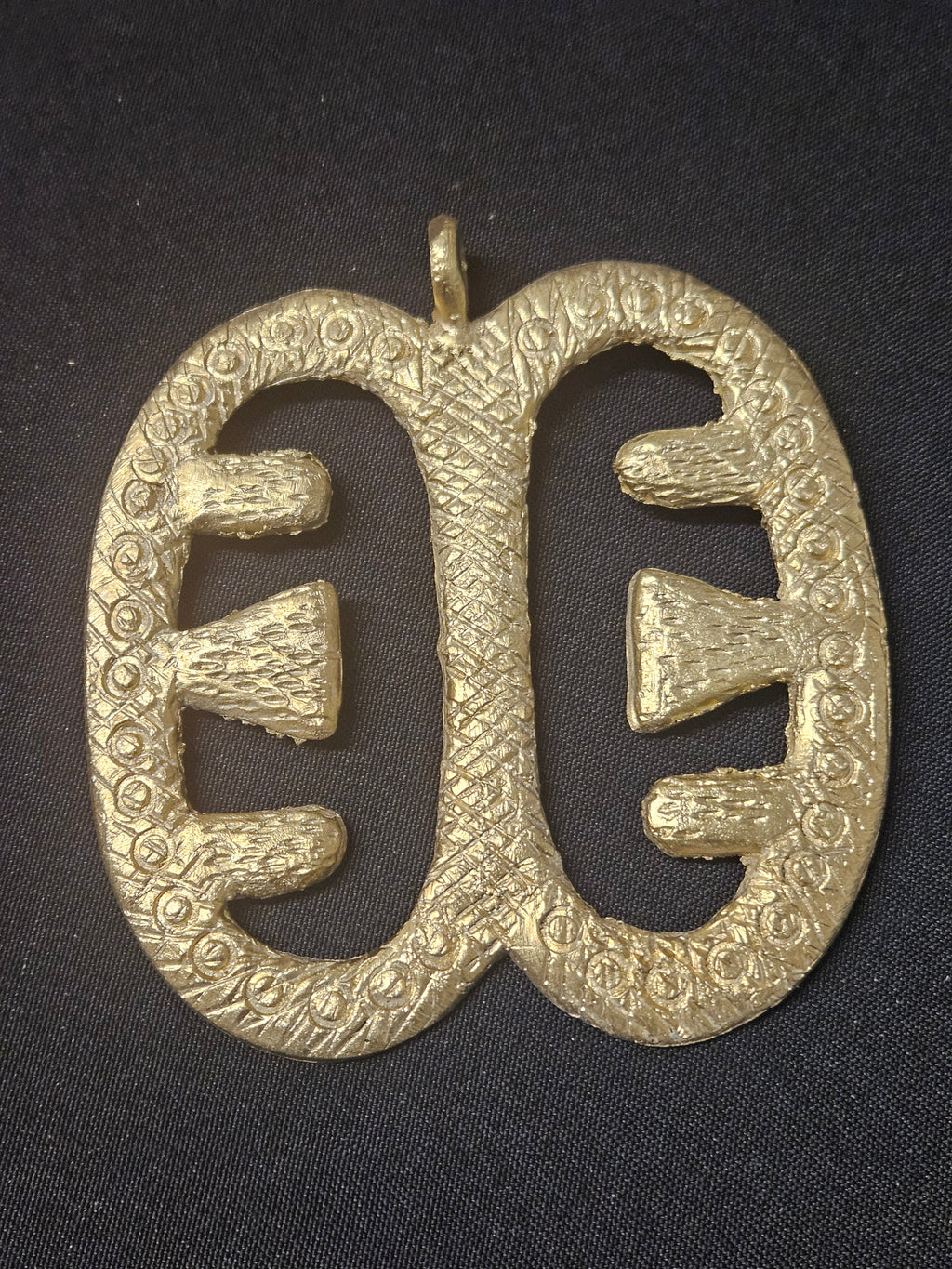 Brass Ese Ne Tekrema pendant with two elongated open sections, inward-facing bar details, a patterned surface, and a top hanging loop on dark fabric.