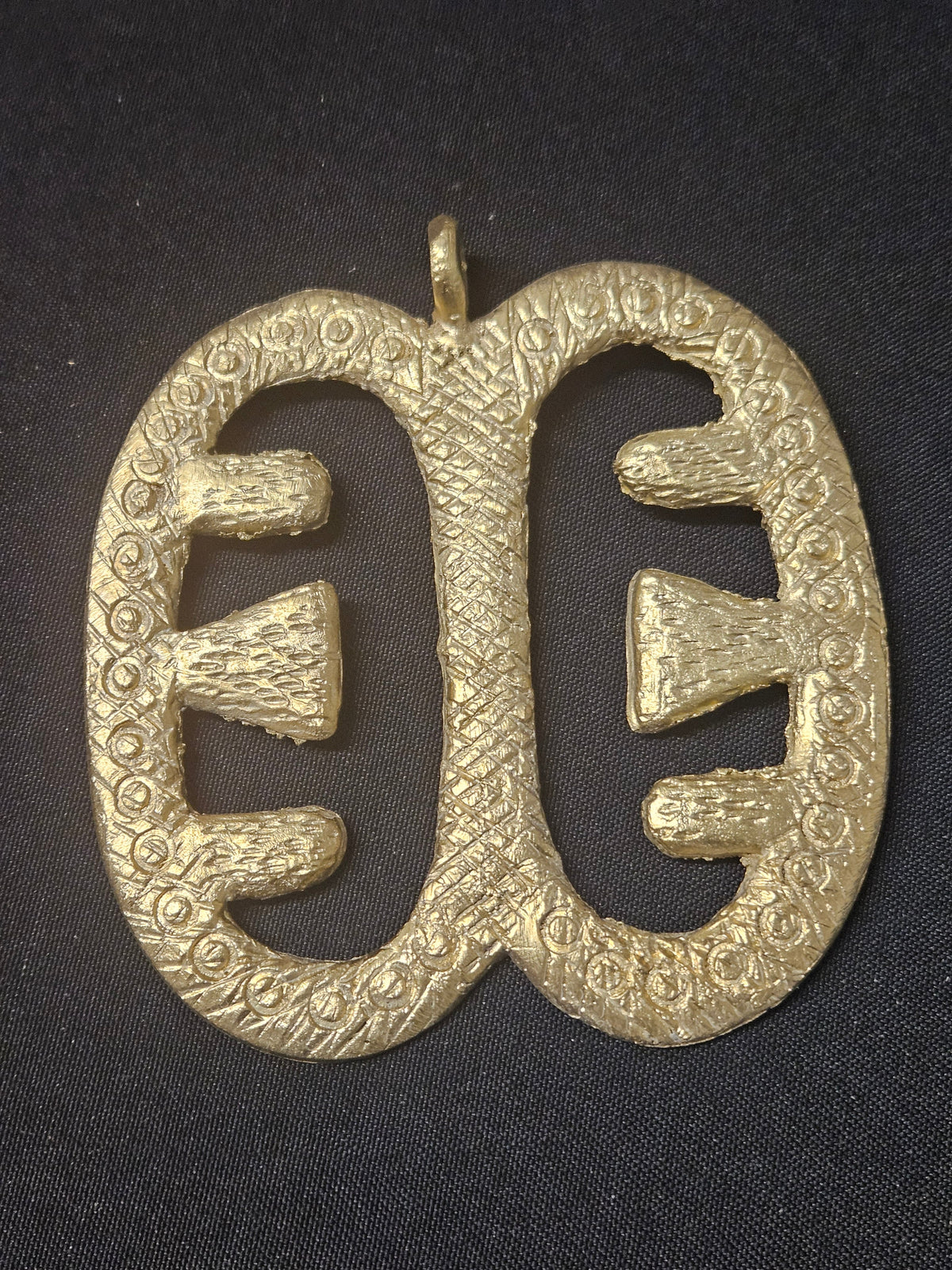 Brass Ese Ne Tekrema pendant with two elongated open sections, inward-facing bar details, a patterned surface, and a top hanging loop on dark fabric.