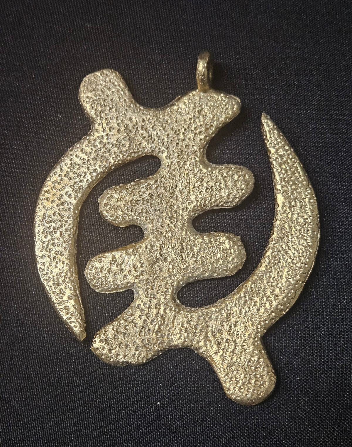 Brass Gye Nyame pendant with curved openwork arms, a textured surface, and a top hanging loop on dark fabric