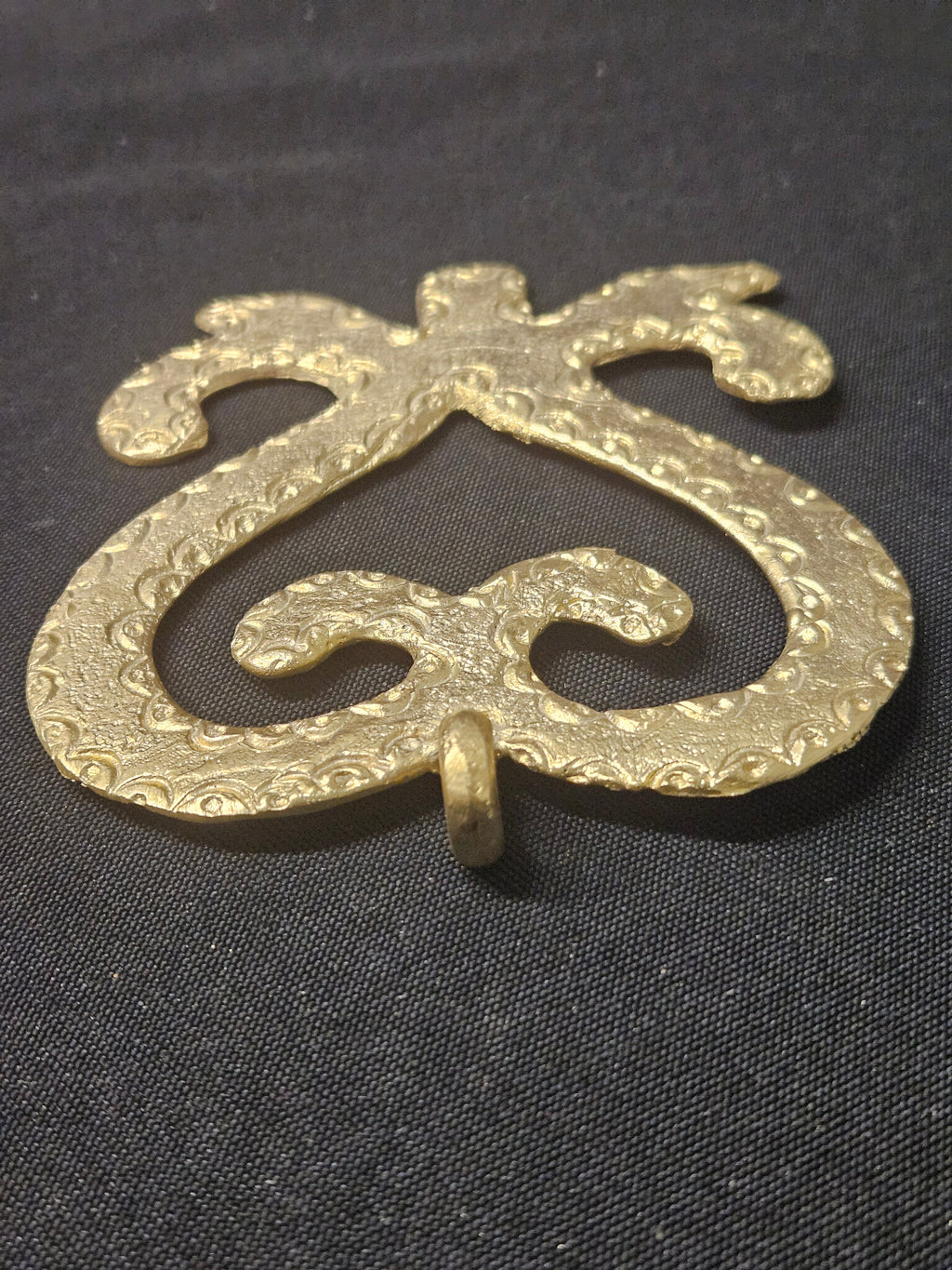Brass heart-shaped Sankofa pendant rotated with its hanging loop at the bottom, showing the open curled design on dark fabric