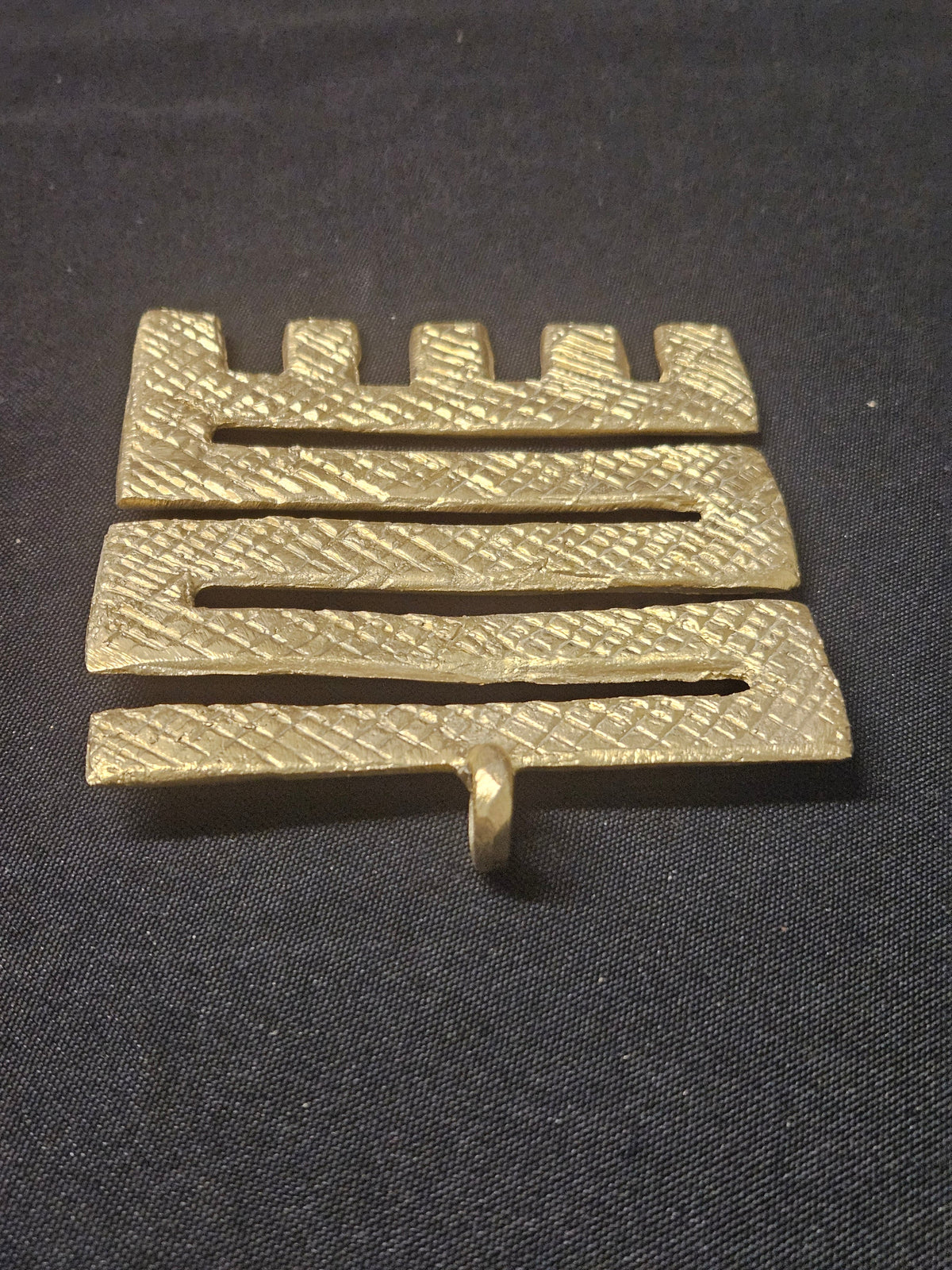 Angled view of the brass Nkyinkyim pendant, rotated with its prongs at the top, showing its zigzag openings and thickness on dark fabric.