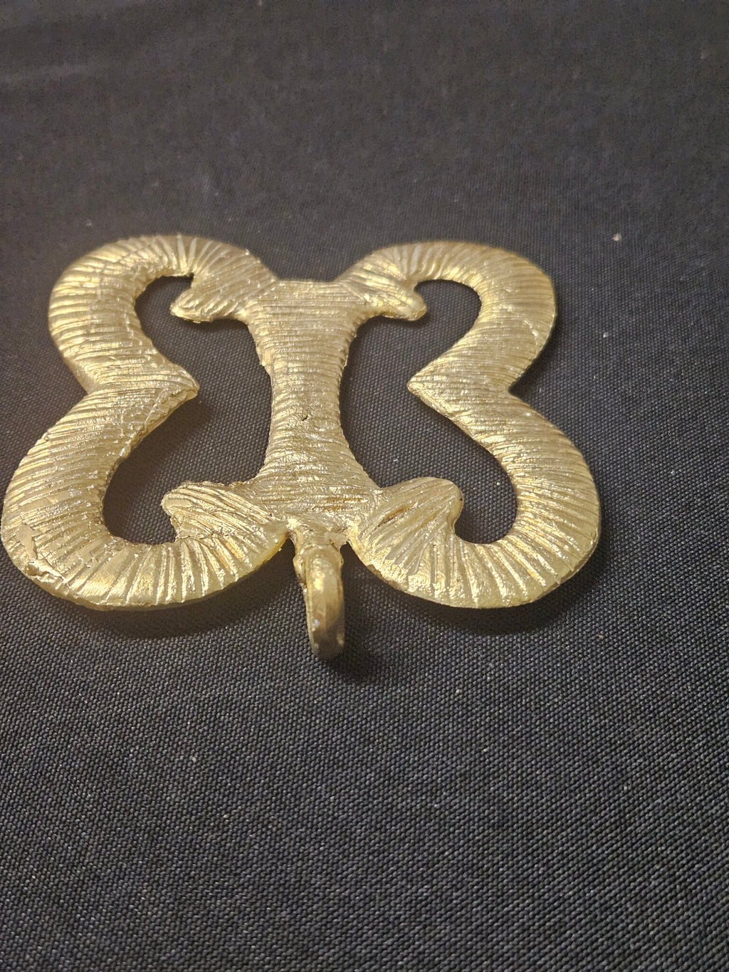 Angled view of the brass Pempamsie pendant showing its curled openwork sections, central connecting bar, and thickness on dark fabric