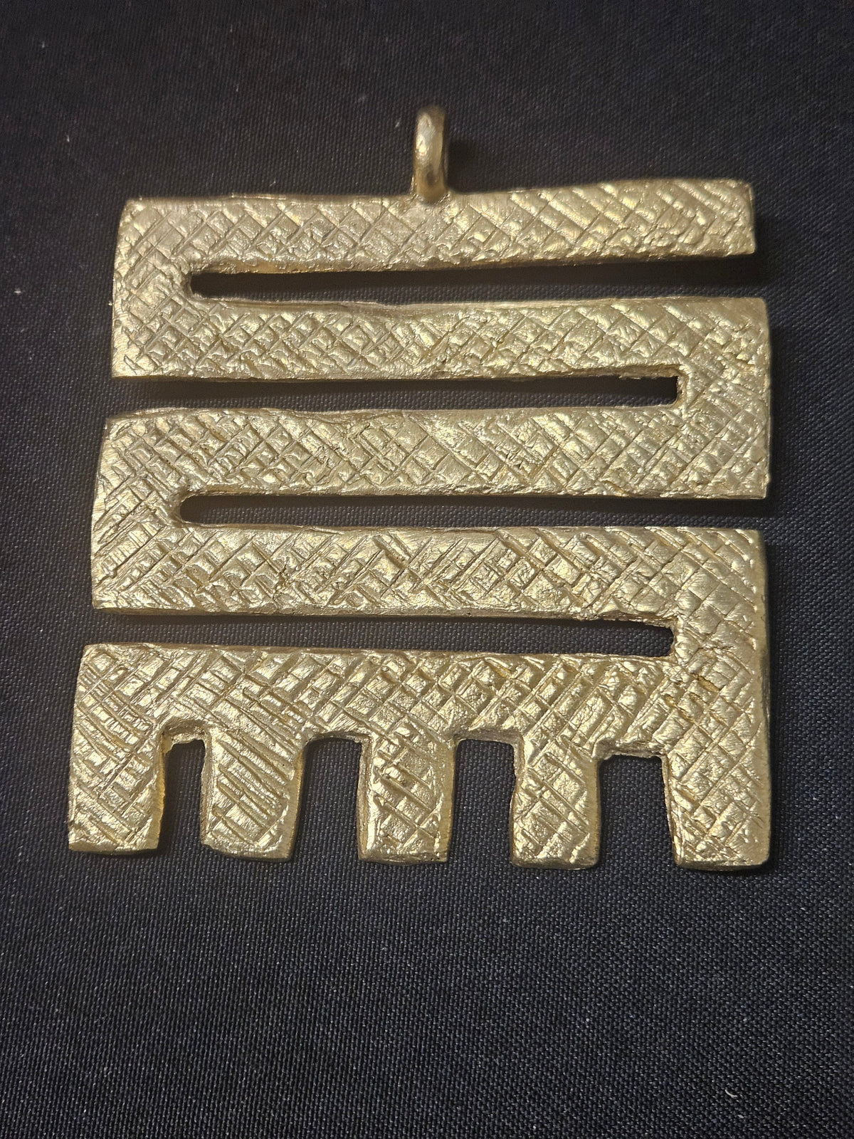 Brass Nkyinkyim pendant with a zigzag openwork design, textured surface patterns, short prongs along the bottom, and a top hanging loop on dark fabric