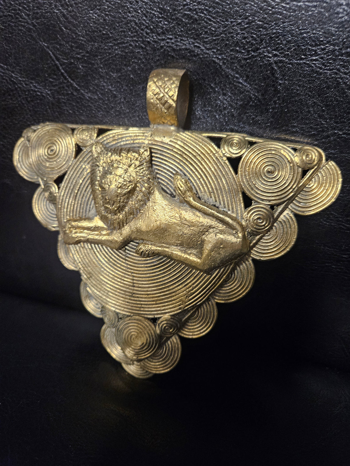Triangular brass pendant with a raised reclining lion over a large spiral disc, smaller spirals around the border, and a textured top hanging loop