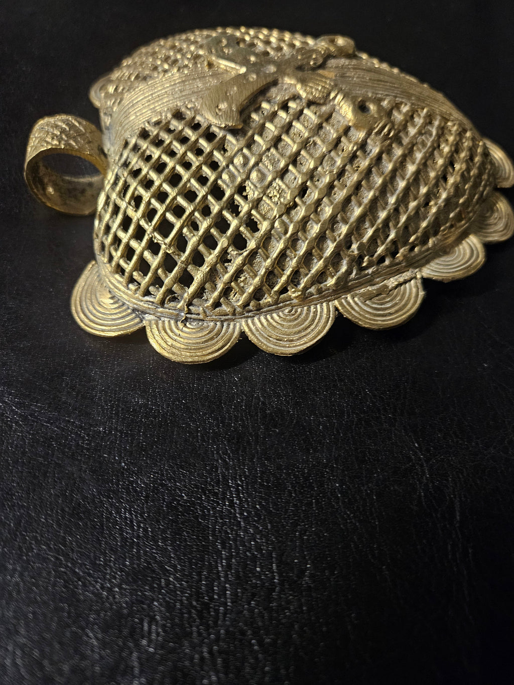 Angled view of the brass Sankofa bird pendant showing its raised bird motif, open diamond lattice, spiral edging, and hanging loop