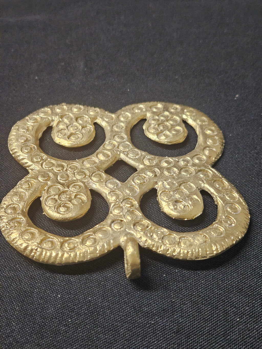 Angled view of the brass Mati Masie pendant showing its four curled openwork sections, patterned surface, and hanging loop on dark fabric