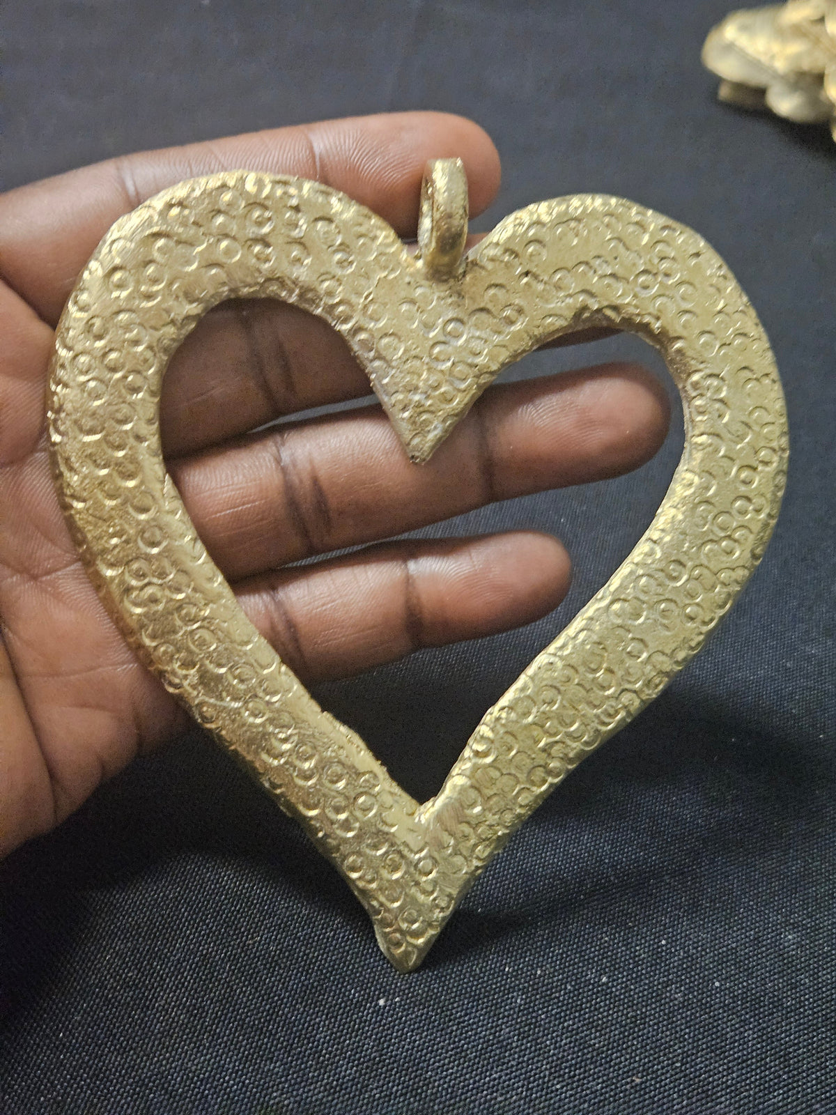 Brass open-heart Akoma pendant held in a hand, showing its size relative to the fingers and patterned surface.