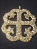 Brass Onyame Dua pendant with four inward-curving open sections around a central cross, patterned surface details, and a top hanging loop on dark fabric