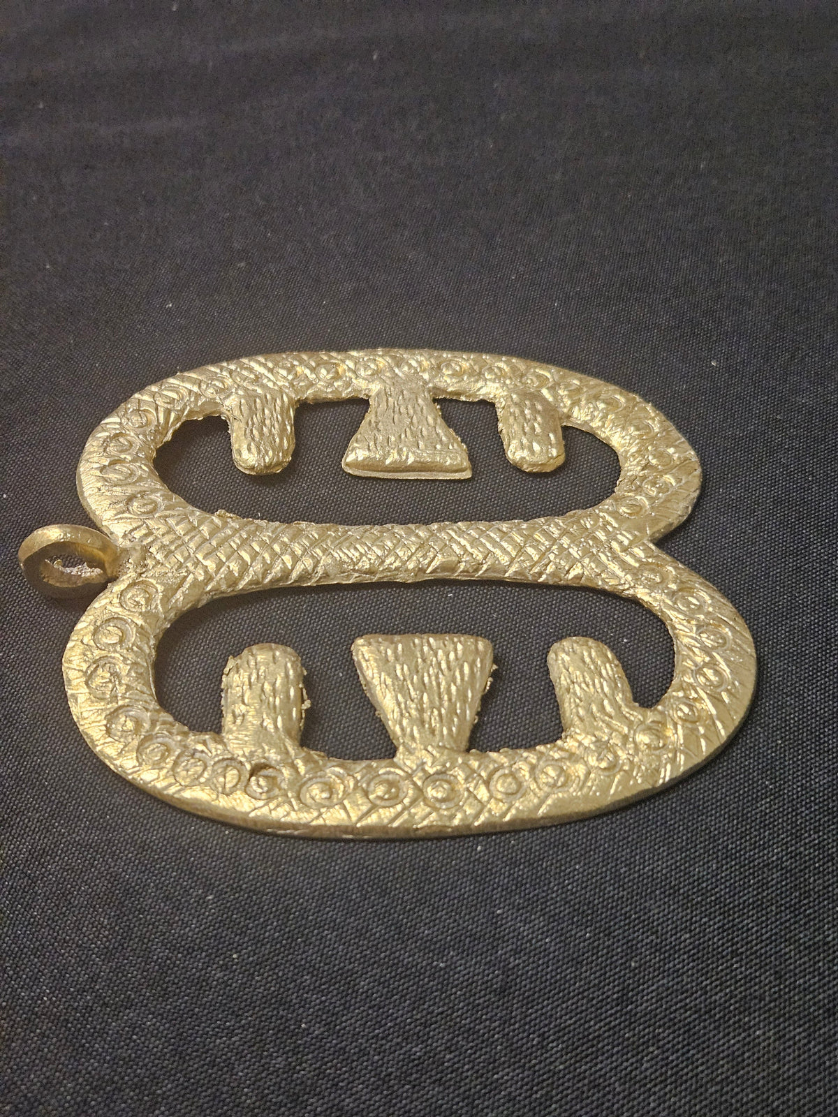 Low angled view of the brass Ese Ne Tekrema pendant lying horizontally, showing its two open sections, inward-facing bars, and thickness on dark fabric