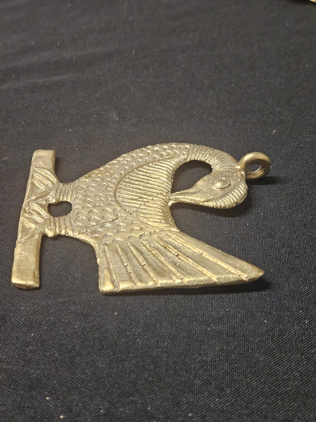 Brass Sankofa bird pendant lying horizontally on dark fabric, showing its bird silhouette, fan-shaped tail, and hanging loop