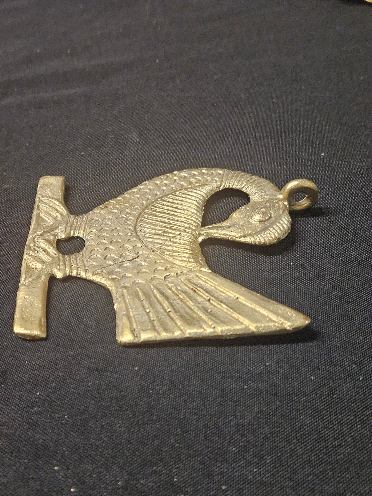 Brass Sankofa bird pendant lying horizontally on dark fabric, showing its bird silhouette, fan-shaped tail, and hanging loop