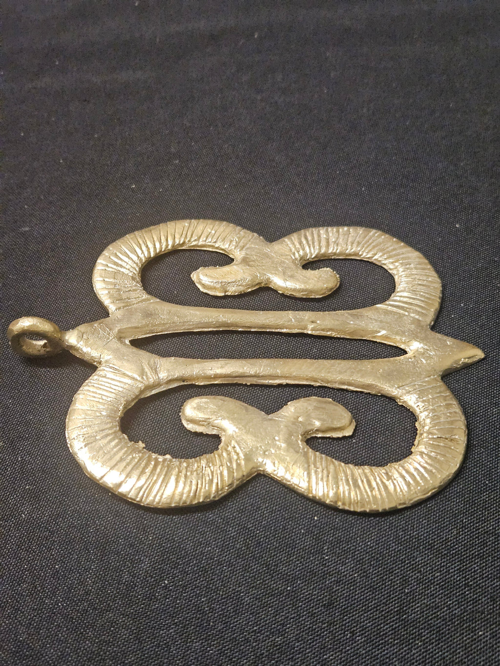 Low angled view of the brass Hye Won Hye pendant lying horizontally, showing its curled openwork design and thickness on dark fabric