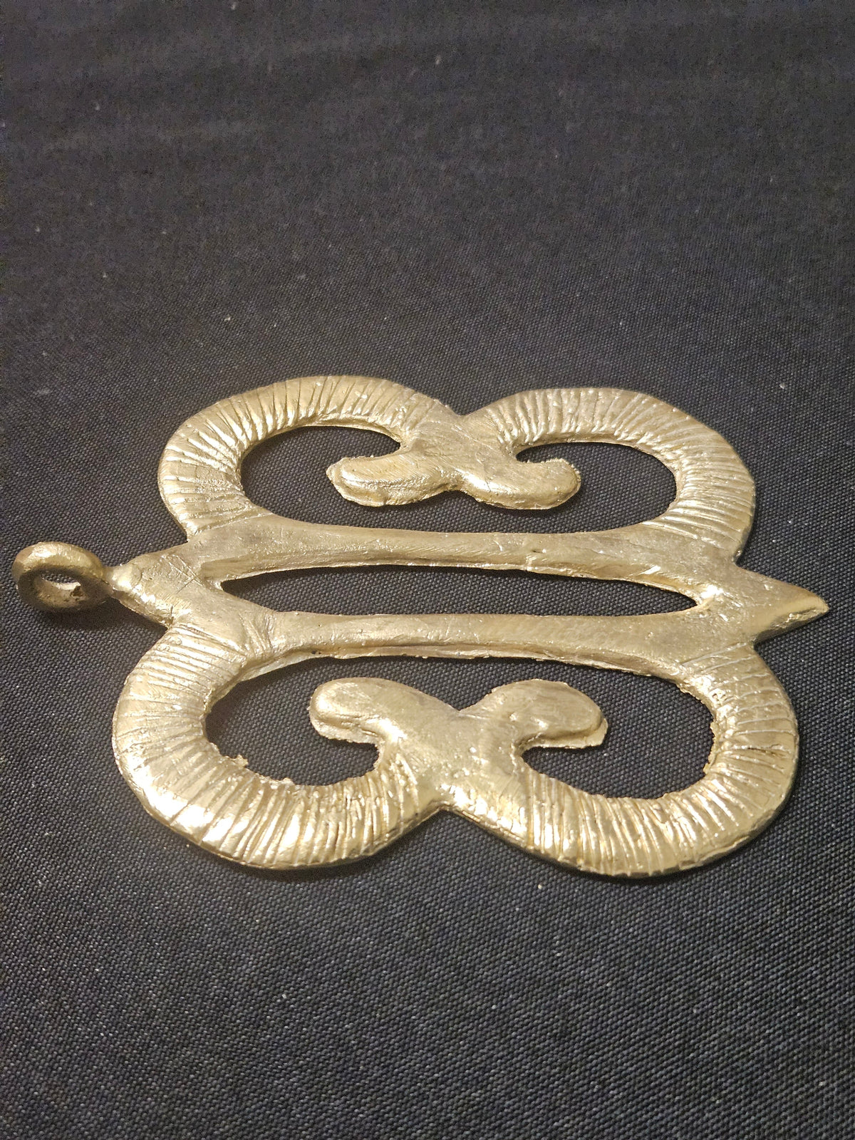 Low angled view of the brass Hye Won Hye pendant lying horizontally, showing its curled openwork design and thickness on dark fabric
