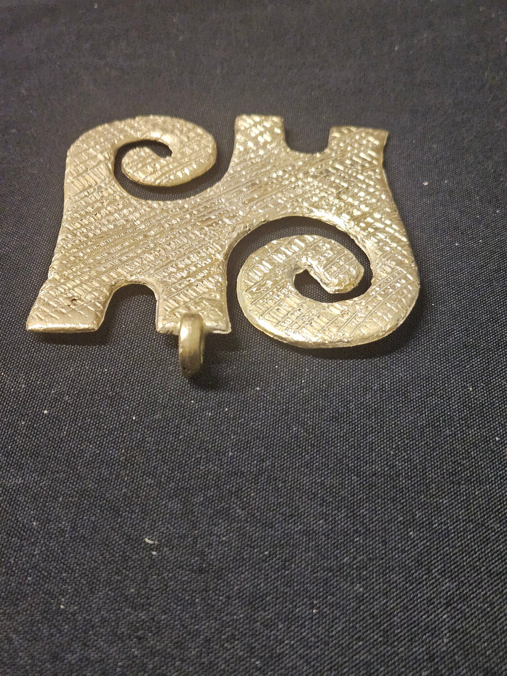 Angled view of the brass Kwaatakye Atiko pendant showing its two spiral curls, edge notches, and thickness on dark fabric