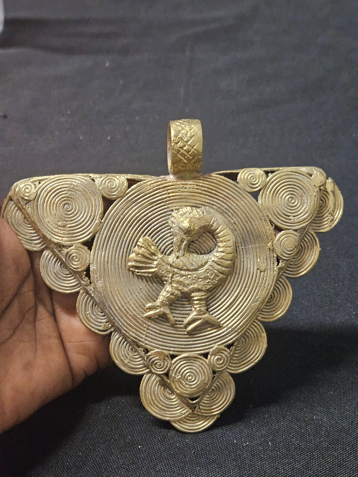 Front of the triangular brass Sankofa bird pendant held in a hand, showing its spiral disc design and hanging loop