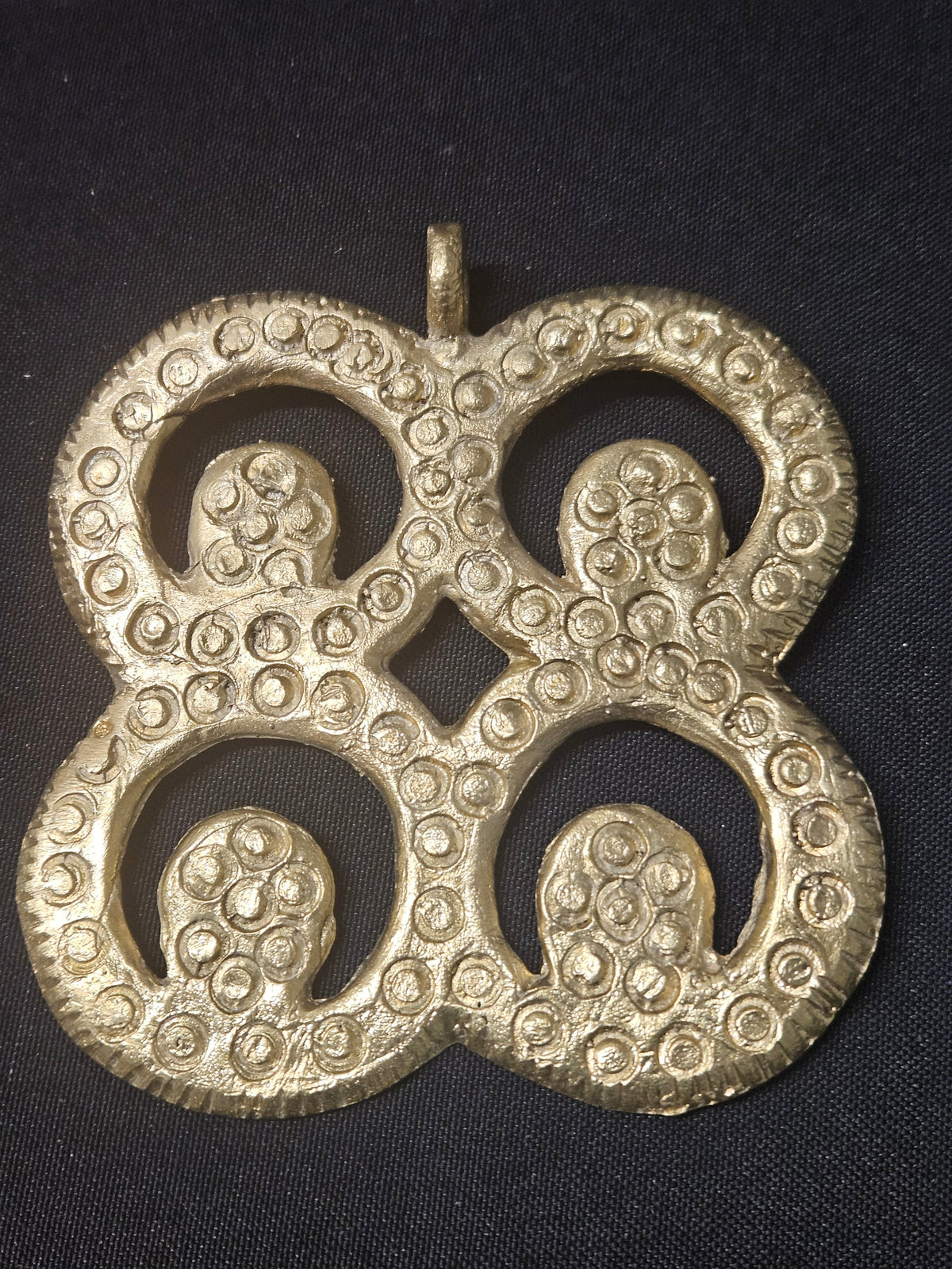 Brass Mati Masie pendant with four circular openwork sections, inward curls, a small diamond-shaped center opening, and a top hanging loop on dark fabric